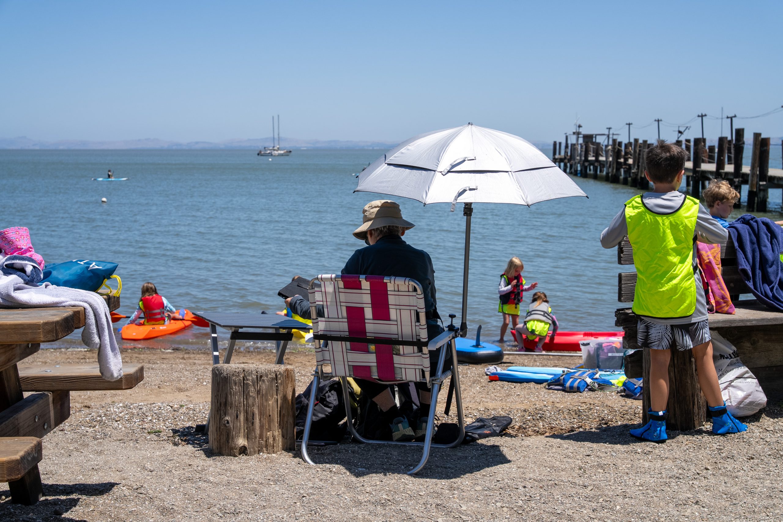 A group gathers at a sunny waterfront, with some sitting under an umbrella and others playing near kayaks and a wooden pier.