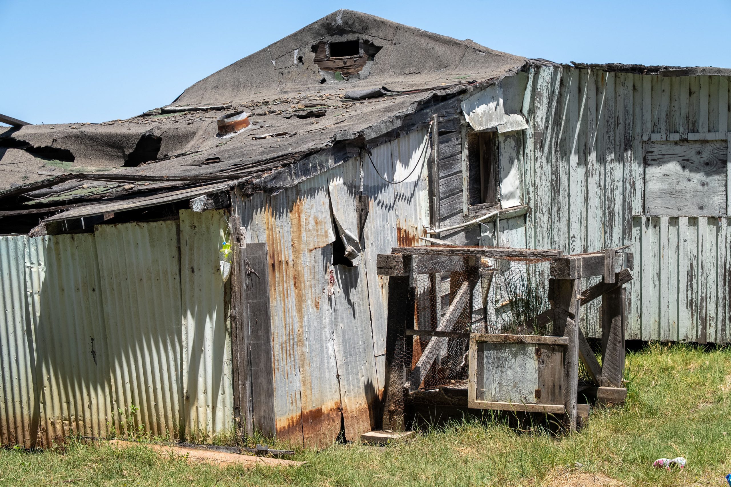 A dilapidated, rusted metal shed with a damaged roof stands in overgrown grass under a clear blue sky.