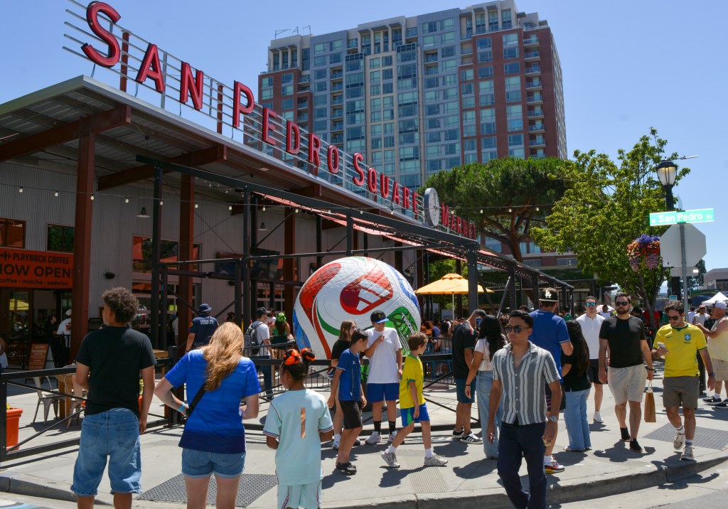 People stroll through San Pedro Square Market on a sunny day, passing a large soccer ball under a prominent red sign.