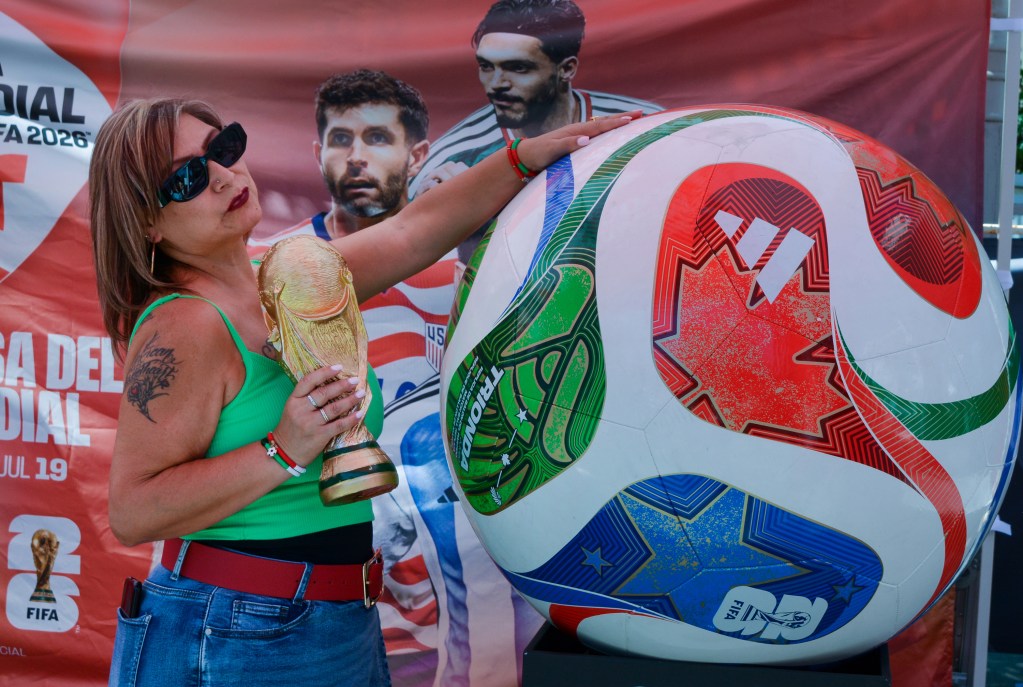 A person holds the FIFA World Cup trophy while touching a large replica of the 2026 World Cup match ball, with promotional banners in the background.