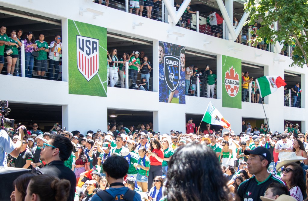 A large crowd of fans in green attire gathers outdoors near a stadium, with banners for the USA and Canada visible on the building's facade.